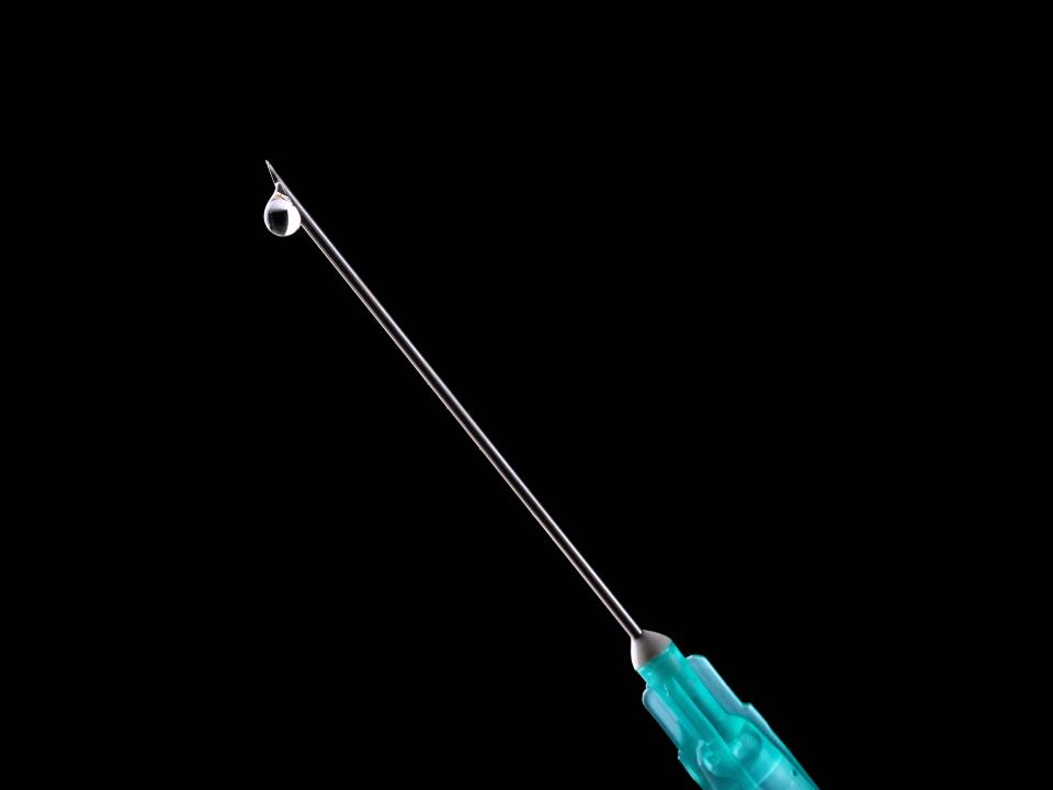 The image shows a teal syringe in front of a black background.  The tip of the needle has a drop of liquid hanging down.
