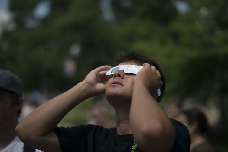 A man looking up at the sky, wearing gray paper eclipse glasses.