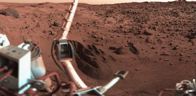 Life on Mars: Part of the robotic arm machine at left, with reddish rocks and sand dunes in the background.